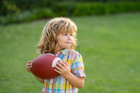 Téléchargez les photos : Portrait d'enfant avec ballon de rugby. Football américain. Enfant prêt à lancer un ballon. Concept sport pour enfants. Activités sportives pour enfants en plein air - en image libre de droit