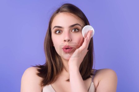 Foto de Beautiful young woman with cotton pad, applying lotion on face, removing makeup. Skin care. Studio portrait of girl cleaning her face with cotton pad, isolated background. Face toner and cotton pad - Imagen libre de derechos