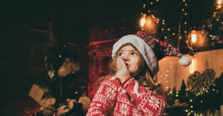 Foto de Feliz Navidad niña está teniendo un secreto. Hermosa niñita con sombrero de Santa. En Navidad. Luz de Navidad - Imagen libre de derechos