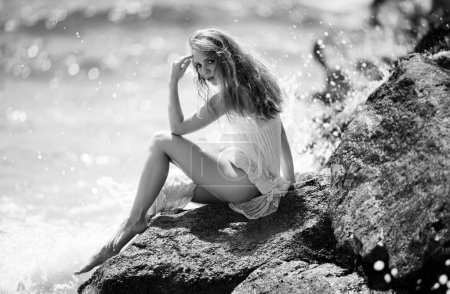 Foto de Vista lateral de una mujer serena sentada en la playa rocosa de arena contra las olas azules. Atractiva mujer joven con vestido sexy de verano en una playa - Imagen libre de derechos