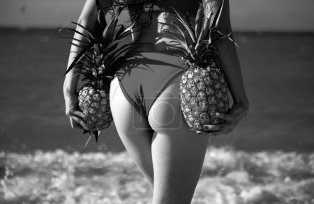 Foto de Mujer sexy sosteniendo piña fresca. Primer plano nalgas de chica en bikini con piña de frutas en el fondo de la playa, vacaciones de verano, comida saludable y fitness - Imagen libre de derechos