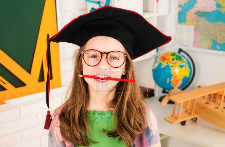 Téléchargez les photos : Joyeux drôle petite fille avec stylo dans la bouche contre tableau noir - en image libre de droit