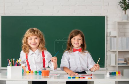 Foto de Niños de la escuela sonriendo niña y niño pintando con pinturas de color y pincel en el aula. Niños felices educación creativa - Imagen libre de derechos