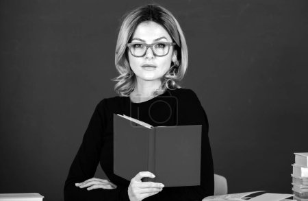 Téléchargez les photos : Modern attractive teacher. Portrait of smart young woman in glasses with book on the blackboard in class at high school or collage. Female student study in university. Study concept - en image libre de droit