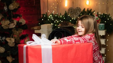 Foto de Feliz Navidad niña quiere abrir un gran regalo de Navidad. Hermosa niña usando suéter rojo. En Navidad. Luz de Navidad - Imagen libre de derechos