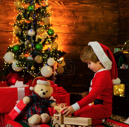 Téléchargez les photos : Petit enfant joue avec des jouets de Noël sur fond d'arbre de Noël. Le gamin attend le Nouvel An. Chapeau de Père Noël. Vacances - en image libre de droit
