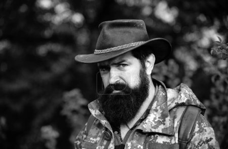 Photo for Portrait of adventure serious man extreme explorer. Man hunter in camouflage outdoor. Brutal hunter, bearded man in the wild forest in the autumn. Portrait man in cowboy hat - Royalty Free Image
