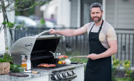 Photo for Men cooking on barbecue grill in yard. Cook at a barbecue grill preparing meat. Guy cooking meat on barbecue for summer family dinner at the backyard of the house - Royalty Free Image