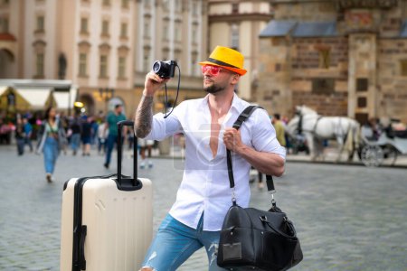 Foto de Hombre viajero con gafas de sol, sombrero y cámara. Viajero masculino con maleta exterior. Retrato de hombre joven caucásico en sombrero y gafas de sol en las vacaciones de verano - Imagen libre de derechos