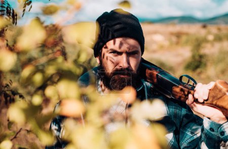 Téléchargez les photos : Un chasseur barbu tenant une arme et marchant dans la forêt. Chasseur avec fusil de chasse en chasse. Gros plan Portrait de hamdsome Hunter - en image libre de droit