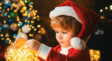 Foto de Niño con luces de guirnalda en el árbol de Navidad en Nochebuena. Es un milagro. Familia con niños celebrando la Navidad en casa. Niño lindo niño alegre humor jugar cerca de árbol de Navidad - Imagen libre de derechos