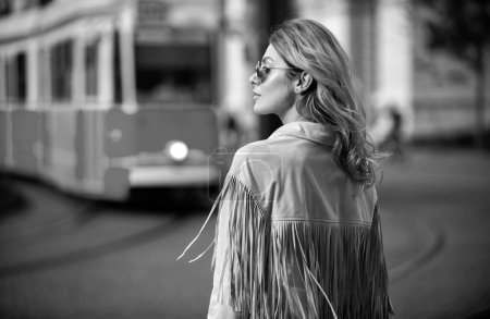 Foto de Foto de estilo callejero de mujer elegante de moda con ropa de moda. Modelo caminando en la calle de la ciudad europea en el fondo del trolebús autobús - Imagen libre de derechos