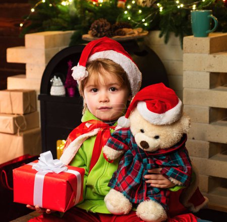Téléchargez les photos : Joyeux petit garçon avec son ours en peluche regarde la caméra. Service de livraison d'hiver pour enfants. Joyeux enfant avec boîte cadeau de Noël. Des petits enfants mignons qui fêtent Noël. Fête de Noël - en image libre de droit