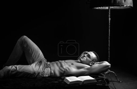 Foto de Hombre guapo en denim azul con libro y torso desnudo. Acuéstate en la cama y relájate. Estilo de vida de lujo - Imagen libre de derechos