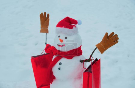 Foto de Feliz invierno. Muñeco de nieve divertido con una zanahoria en lugar de una nariz y en un sombrero de punto caliente en un prado cubierto de nieve sobre un fondo de nieve borrosa - Imagen libre de derechos