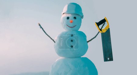 Téléchargez les photos : Hiver constructeur de bonhomme de neige dans casque tenir scie. Bonhomme de neige sur le fond extérieur de neige. Bannière de Noël avec bonhomme de neige. Bonhomme de neige prêt pour les travaux de construction et de réparation - en image libre de droit