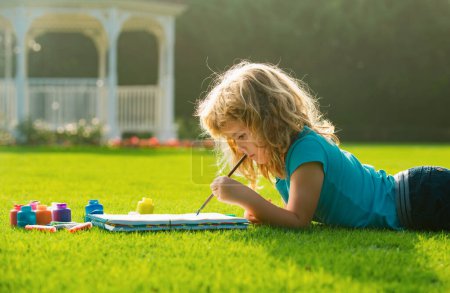 Téléchargez les photos : Enfants peignant dans la nature printanière. Kid dessine dans le parc posé dans l'herbe en s'amusant sur fond de nature. Enfant art, créativité enfant concept - en image libre de droit