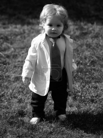 Téléchargez les photos : Bébé enfant sur l'herbe verte dans le parc d'été. Petit homme drôle en costume, veste et cravate - en image libre de droit