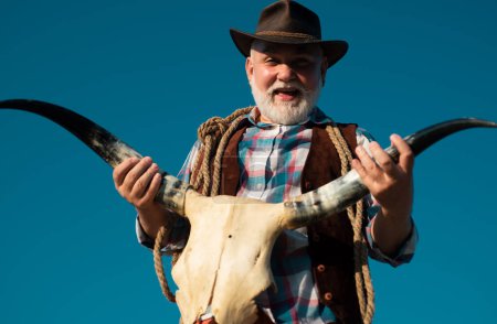 Foto de Viejo vaquero salvaje del oeste con cráneo de vaca. Barbudo hombre occidental con chaqueta marrón y sombrero - Imagen libre de derechos