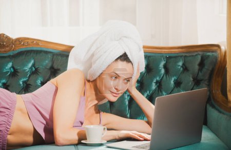 Téléchargez les photos : Attrayant jeune fille étudiante en pyjama et serviettes étudier à la maison, à l'aide d'un ordinateur portable, boire du café - en image libre de droit