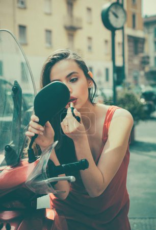 Téléchargez les photos : Femme qui se maquille dans la rue. Modèle sexy fille de mode avec lèvres rouges mettre rouge à lèvres regardant dans une moto miroir - en image libre de droit