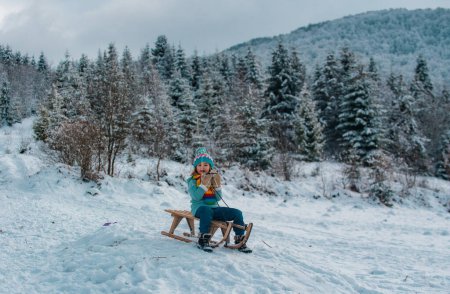 Téléchargez les photos : Garçon luge sur la montagne d'hiver, profitant d'une balade en traîneau dans un magnifique parc d'hiver enneigé. Scène hivernale avec forêt enneigée - en image libre de droit