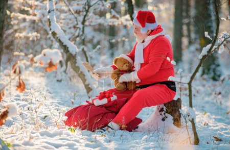 Photo for Santa with Gift and teddy bear read wish list in the winter field. Santa Claus on Christmas Eve is carrying presents to children in a bag - Royalty Free Image