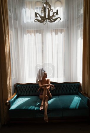 Foto de Niña descansando y acostada en un acogedor sofá cama en el dormitorio de lujo una mañana fácil. Atractiva mujer sexy que se relaja en la habitación del hotel, con albornoces y toallas, beber café - Imagen libre de derechos