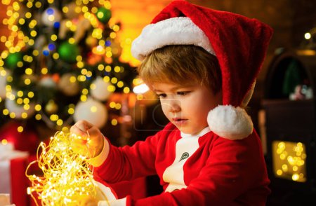Foto de Lindo niño de Santa con luces de guirnalda de Navidad. Feliz bebé de santa con regalos de año nuevo en el fondo navideño. Niño pequeño decorando árbol de Navidad y regalos de apertura - Imagen libre de derechos