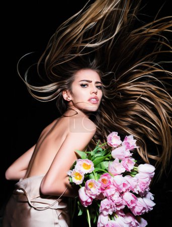 Téléchargez les photos : Jeune femme avec les fleurs et les cheveux longs. Publicité de fleuriste. Salon de beauté. Coiffure de printemps. Cheveux longs en mouvement - en image libre de droit