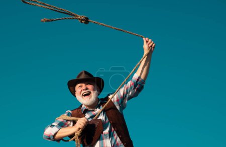 Foto de Viejo vaquero salvaje del oeste con cuerda de lazo. Hombre occidental barbudo con chaqueta marrón y sombrero de captura de caballo o vaca - Imagen libre de derechos