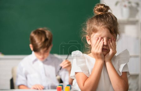 Téléchargez les photos : Une écolière intelligente pleure. Écolière en uniforme couvrant son visage avec son bras pleurant triste d'intimidation à l'école debout devant le tableau noir. Concept de problème de l'enfance et de l'éducation - en image libre de droit