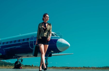 Téléchargez les photos : Aéroport et hôtesse de l'air charmante sur fond d'avion bleu. Portrait de charmante hôtesse en uniforme bleu. Femme et avion commercial. Toute la longueur de l'hôtesse de l'air debout par avion à réaction - en image libre de droit