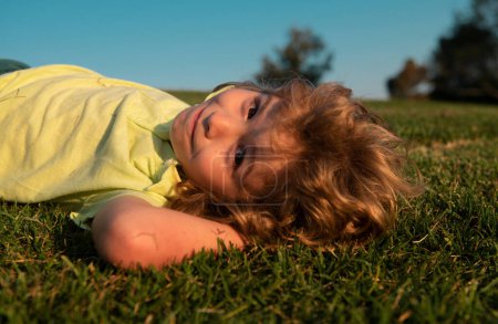 Téléchargez les photos : Portrait d'un enfant souriant couché sur de l'herbe verte. Petit garçon mignon profitant de la nature en plein air. Enfant en bonne santé jouant dehors dans le parc d'été - en image libre de droit