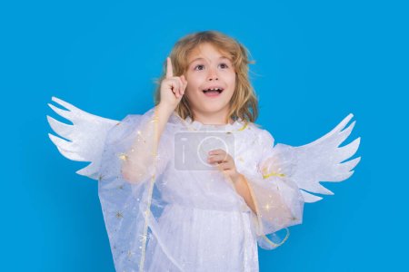 Foto de Ángel niño apuntando hacia arriba, gesto puntual. Día de San Valentín. Rubia linda niña con alas de ángel en un fondo de estudio azul. Niño ángel feliz - Imagen libre de derechos