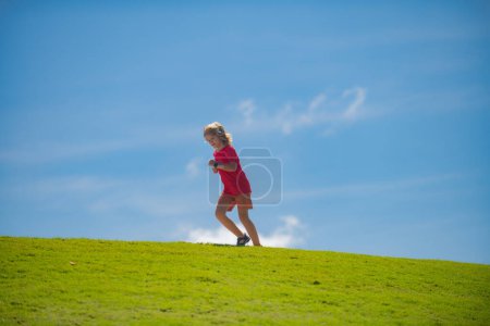 Foto de Lindo chico corriendo sobre hierba verde y cielo azul. Carrera y deporte saludable para los niños. Niño corriendo en el parque de verano. Ejecutar entrenamiento al aire libre. Niños deportistas - Imagen libre de derechos