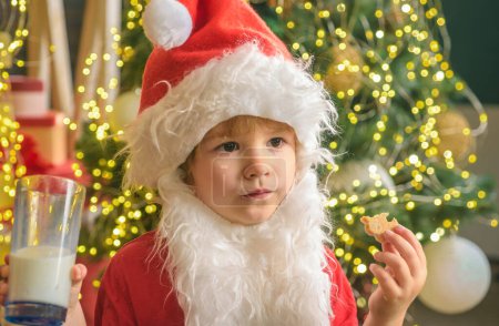 Foto de Niño Santa Claus disfrutando en servido pastel de jengibre y leche. Niño Santa Claus toma una galleta en Nochebuena como un regalo de agradecimiento por dejar regalos a un niño o niña agradecidos - Imagen libre de derechos