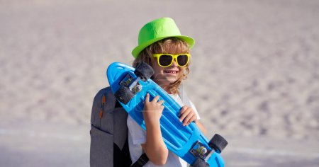 Téléchargez les photos : Journée des enfants. Joyeux enfant élégant dans les lunettes de soleil et chapeau d'été de mode posant avec planche à roulettes en plein air - en image libre de droit