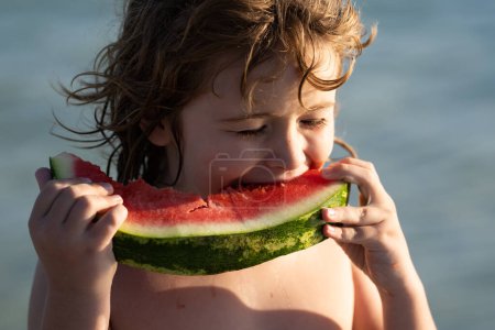 Photo for Close up summer portrait of cute child play at sea beach and eat watermelon. Little boy biting watermelon on beach. Kid is picking watermelon outdoor. Kids funny face and watermelon, closeup - Royalty Free Image
