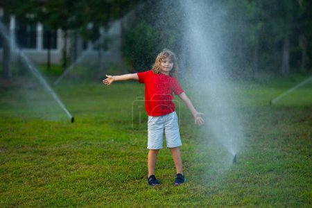 Téléchargez les photos : Child play near automatic sprayers in the garden. Watering in the garden. Kid freshness of nature. Automatic lawn sprinkler watering grass. Garden irrigation system watering lawn. Sprinkler system - en image libre de droit