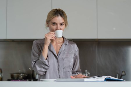Téléchargez les photos : Femme sexy assis dans la cuisine le matin et boire du café. Sensuelle femme au foyer lecture livre ou magazine - en image libre de droit