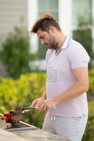 Téléchargez les photos : Homme cuisinant de la viande sur le barbecue dans la cour de la maison. Un bel homme qui prépare un barbecue. Chef cuisinier de barbecue. Cuisiner en préparant de délicieux barbecue grillé, viande barbecue. Grill et barbecue - en image libre de droit