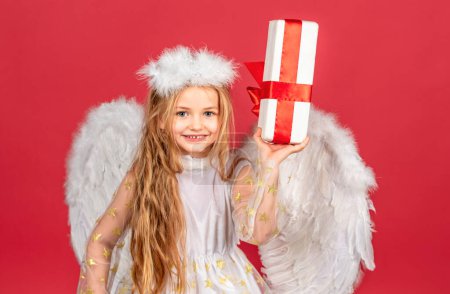 Photo for Valentine. Happy smiling cupid girl. Kid angel with present. Little angel with white wings holds gift. Playful angelic little girl. Cute angel child girl with angels wings, isolated on red. Valentines - Royalty Free Image