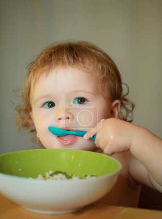 Téléchargez les photos : Portrait d'un drôle de petit garçon mangeant dans une assiette tenant une cuillère en gros plan. Une alimentation saine pour les enfants - en image libre de droit