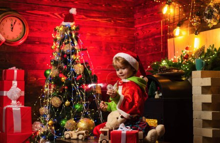 Foto de El pequeño Santa está jugando con juguetes junto al árbol de Navidad. Niño pequeño lleva ropa de Santa Claus - Imagen libre de derechos
