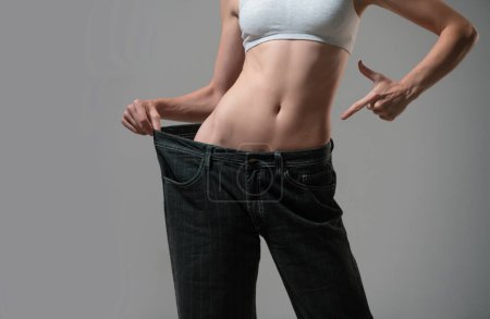 Téléchargez les photos : Concept de régime avec un jean surdimensionné. Femme maigre en trop gros jeans. Concept de perte de poids réussie. Mince fille en gros pantalon sur un fond gris - en image libre de droit