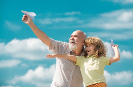 Téléchargez les photos : Jeune petit-fils et vieux grand-père avec avion en papier jouet sur fond de ciel d'été. Enfant garçon avec des rêves de vol - en image libre de droit