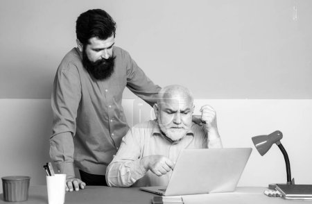 Foto de Dos generaciones diferentes sentados en el escritorio con ordenadores portátiles. Hijo adulto enseñando a papá mayor maduro a usar la computadora. Hombre mayor enseñando portátil a su hijo mayor - Imagen libre de derechos