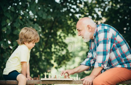 Téléchargez les photos : Petit garçon jouant aux échecs avec son grand-père. Pièce d'échecs. Senior pense à son prochain coup dans un jeu d'échecs. Grand-père jouant aux échecs avec son petit-fils - en image libre de droit