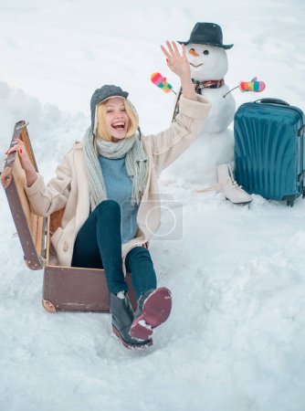 Foto de Chica joven feliz jugando en nieve fresca y haciendo muñeco de nieve en el hermoso día de invierno soleado al aire libre en la naturaleza. Concepto de viaje de invierno. Vacaciones de invierno. Maleta de trineo - Imagen libre de derechos